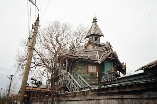 A rustic wooden house with intricate architectural details and a steep roof, surrounded by bare trees and power lines. The structure appears old and weathered, lending a historical or vintage feel.