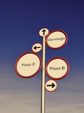 A tall metal signpost features multiple circular signs with red borders against a clear blue sky. Each sign provides directional information: 'Dermaga' with an upwards arrow, 'Plaza D' with a left arrow, and 'Plaza B' with a right arrow.