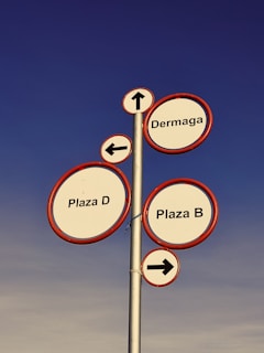 A tall metal signpost features multiple circular signs with red borders against a clear blue sky. Each sign provides directional information: 'Dermaga' with an upwards arrow, 'Plaza D' with a left arrow, and 'Plaza B' with a right arrow.