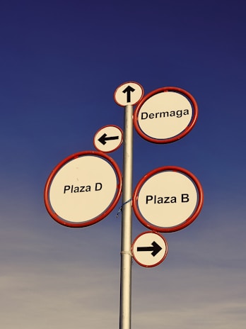A tall metal signpost features multiple circular signs with red borders against a clear blue sky. Each sign provides directional information: 'Dermaga' with an upwards arrow, 'Plaza D' with a left arrow, and 'Plaza B' with a right arrow.