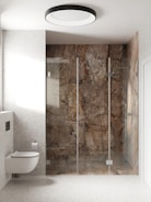 A modern bathroom with a wall-mounted toilet and a large walk-in shower. The shower has glass doors and is enclosed by walls with a unique brown and gray marble design. The bathroom is bright with off-white speckled walls and a matching floor. A circular ceiling light provides illumination.