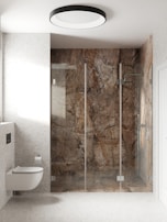 A modern bathroom with a wall-mounted toilet and a large walk-in shower. The shower has glass doors and is enclosed by walls with a unique brown and gray marble design. The bathroom is bright with off-white speckled walls and a matching floor. A circular ceiling light provides illumination.