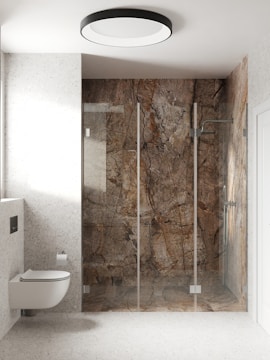 A modern bathroom with a wall-mounted toilet and a large walk-in shower. The shower has glass doors and is enclosed by walls with a unique brown and gray marble design. The bathroom is bright with off-white speckled walls and a matching floor. A circular ceiling light provides illumination.