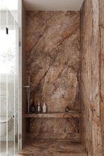 A luxurious bathroom shower area featuring elegant stone walls and floors with pronounced natural patterns. The walls have a rich, earthy appearance with dark and light brown streaks. Within the shower, several modern bottles and a brush are neatly placed on a shelf. The showerhead and controls appear sleek and contemporary. Adjacent to the shower is a glimpse of a toilet with a textured wall and frosted glass door.