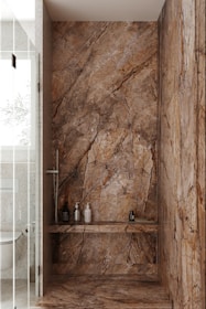 A luxurious bathroom shower area featuring elegant stone walls and floors with pronounced natural patterns. The walls have a rich, earthy appearance with dark and light brown streaks. Within the shower, several modern bottles and a brush are neatly placed on a shelf. The showerhead and controls appear sleek and contemporary. Adjacent to the shower is a glimpse of a toilet with a textured wall and frosted glass door.