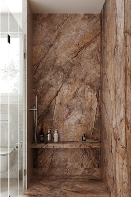 A luxurious bathroom shower area featuring elegant stone walls and floors with pronounced natural patterns. The walls have a rich, earthy appearance with dark and light brown streaks. Within the shower, several modern bottles and a brush are neatly placed on a shelf. The showerhead and controls appear sleek and contemporary. Adjacent to the shower is a glimpse of a toilet with a textured wall and frosted glass door.