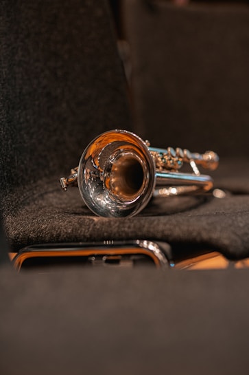 A shiny brass trumpet is resting on a dark, textured surface, likely a seat in a concert hall. The instrument's bell is prominently visible, reflecting the warm ambient light.