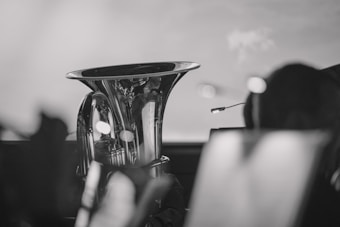 A polished brass instrument, possibly a tuba or euphonium, is prominently featured at the center. The background is soft and blurred, suggesting a performance or practice setting with music stands and other musicians. The image is in black and white, giving it a classic and timeless appearance.