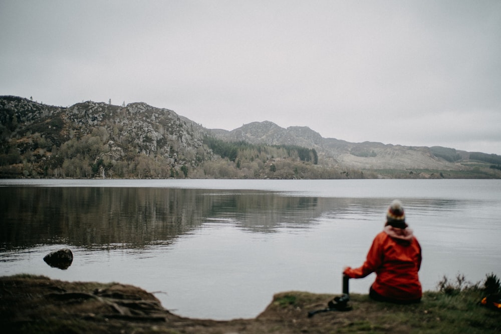 a man sitting on a rock by a lake with mountains in the background