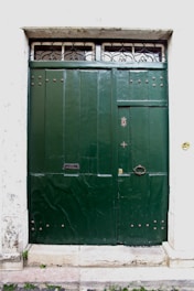 A large, dark green double door made of wood or metal with decorative rivets and a small, round handle. Above the door, there is a narrow transom window featuring ornate iron grillwork. The surrounding wall is white and textured, with some signs of wear and age.