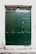 A large, dark green double door made of wood or metal with decorative rivets and a small, round handle. Above the door, there is a narrow transom window featuring ornate iron grillwork. The surrounding wall is white and textured, with some signs of wear and age.