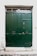 A large, dark green double door made of wood or metal with decorative rivets and a small, round handle. Above the door, there is a narrow transom window featuring ornate iron grillwork. The surrounding wall is white and textured, with some signs of wear and age.