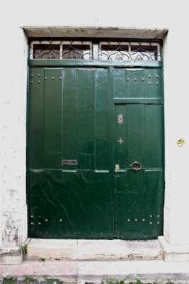 A large, dark green double door made of wood or metal with decorative rivets and a small, round handle. Above the door, there is a narrow transom window featuring ornate iron grillwork. The surrounding wall is white and textured, with some signs of wear and age.