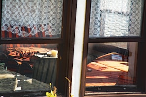 A cozy interior seen through a window with lace curtains. Inside, a warm-toned room features a small table with two candles and a striped armchair. Sunlight filters through, casting shadows and highlighting the orange and brown tones of the furniture.