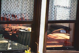 A cozy interior seen through a window with lace curtains. Inside, a warm-toned room features a small table with two candles and a striped armchair. Sunlight filters through, casting shadows and highlighting the orange and brown tones of the furniture.