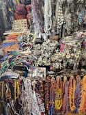 A vibrant and eclectic assortment of colorful beads, necklaces, and various jewelry items displayed in a market setting. The collection includes intricate designs with a mix of beads, chains, and gemstones. The background shows a variety of other accessories and possibly some symbolic or traditional items, creating a rich, textured scene.