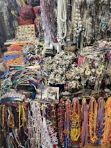 A vibrant and eclectic assortment of colorful beads, necklaces, and various jewelry items displayed in a market setting. The collection includes intricate designs with a mix of beads, chains, and gemstones. The background shows a variety of other accessories and possibly some symbolic or traditional items, creating a rich, textured scene.