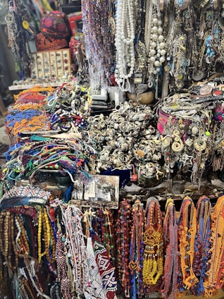A vibrant and eclectic assortment of colorful beads, necklaces, and various jewelry items displayed in a market setting. The collection includes intricate designs with a mix of beads, chains, and gemstones. The background shows a variety of other accessories and possibly some symbolic or traditional items, creating a rich, textured scene.