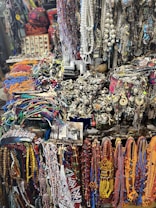 A vibrant and eclectic assortment of colorful beads, necklaces, and various jewelry items displayed in a market setting. The collection includes intricate designs with a mix of beads, chains, and gemstones. The background shows a variety of other accessories and possibly some symbolic or traditional items, creating a rich, textured scene.