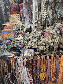 A vibrant and eclectic assortment of colorful beads, necklaces, and various jewelry items displayed in a market setting. The collection includes intricate designs with a mix of beads, chains, and gemstones. The background shows a variety of other accessories and possibly some symbolic or traditional items, creating a rich, textured scene.