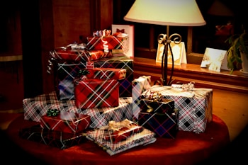 A collection of gift boxes wrapped in various plaid and striped patterns is stacked on a table. A warm lamp illuminates the scene, creating a cozy and inviting atmosphere. Greeting cards are placed in the background near a window, adding to the festive setting.
