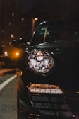 A close-up view of a car headlight at night, with the light on and glowing against the dark background. The sleek design is partially visible, showing reflections on the shiny black surface. Streetlights and building windows softly illuminate the surroundings.