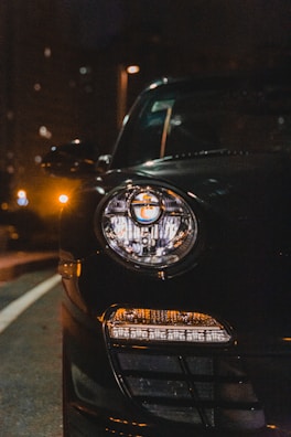 A close-up view of a car headlight at night, with the light on and glowing against the dark background. The sleek design is partially visible, showing reflections on the shiny black surface. Streetlights and building windows softly illuminate the surroundings.