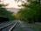 A serene landscape depicting an old railway track surrounded by lush green trees. The tracks lead towards a distant view of hills under a pale sky. A few people are walking along the tracks, adding a sense of scale and activity to the tranquil setting.