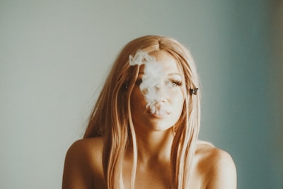 A person with long hair is exhaling a cloud of smoke indoors. Their face is partially obscured by the smoke, and they have bare shoulders with a neutral background behind them.