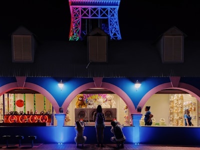 A nighttime scene of a brightly lit, colorful carnival game booth with an illuminated structure resembling a small Eiffel Tower in the background. The booth has three archways; the left displays a game with red and green targets, the center showcases several stuffed animals as prizes, and the right has two people in blue uniforms. Two individuals are seen interacting with the games and prizes.