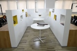 A modern office space with two round white tables, one positioned higher than the other. Books are placed on the tables, and surrounding the area are white walls with rectangular cutouts painted in a contrasting yellow color, displaying various items. The office features wooden flooring and several black office chairs visible in the background.