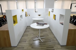 A modern office space with two round white tables, one positioned higher than the other. Books are placed on the tables, and surrounding the area are white walls with rectangular cutouts painted in a contrasting yellow color, displaying various items. The office features wooden flooring and several black office chairs visible in the background.