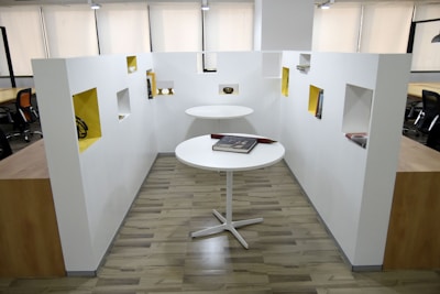 A modern office space with two round white tables, one positioned higher than the other. Books are placed on the tables, and surrounding the area are white walls with rectangular cutouts painted in a contrasting yellow color, displaying various items. The office features wooden flooring and several black office chairs visible in the background.