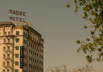 A historic multi-story hotel building with prominent signage reading 'Padre Hotel' sits under a clear sky. The architecture features classic design elements, and a fire escape is visible on the side. Leafy trees frame the scene, adding a natural element to the urban setting.