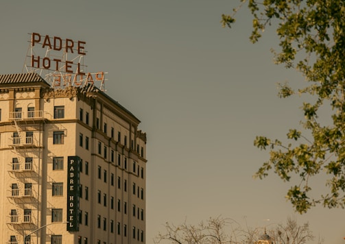 A historic multi-story hotel building with prominent signage reading 'Padre Hotel' sits under a clear sky. The architecture features classic design elements, and a fire escape is visible on the side. Leafy trees frame the scene, adding a natural element to the urban setting.