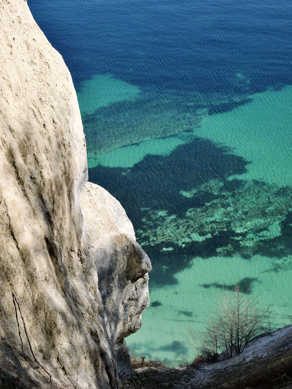 The image depicts a breathtaking view of a cliff overlooking a serene body of water, possibly an ocean or sea. The cliff is white and jagged, with patches of green moss growing on it. The water below mirrors the sky above in a beautiful shade of blue-green, creating a stunning contrast between the rocky cliff and the calm water surface. A small boat can be seen floating near the shoreline, adding to the overall tranquility of the scene.