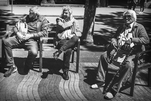 Three elderly people are seated on benches in an outdoor setting with dappled sunlight creating patterns of light and shadow. The individuals are dressed in casual attire, and they appear relaxed and engaged in their thoughts. The scene conveys a sense of leisurely time in a public space.