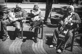 Three elderly people are seated on benches in an outdoor setting with dappled sunlight creating patterns of light and shadow. The individuals are dressed in casual attire, and they appear relaxed and engaged in their thoughts. The scene conveys a sense of leisurely time in a public space.