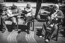 Three elderly people are seated on benches in an outdoor setting with dappled sunlight creating patterns of light and shadow. The individuals are dressed in casual attire, and they appear relaxed and engaged in their thoughts. The scene conveys a sense of leisurely time in a public space.