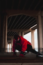 A person dressed in a red and black superhero costume is crouched on the ground under a large overpass. The setting is urban and dimly lit, with large concrete pillars and beams forming the background.