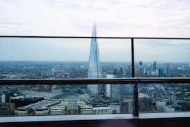 A cityscape featuring a prominent glass skyscraper rising high above the surrounding buildings. The view includes several modern high-rise structures against a backdrop of a vast urban landscape. A railing in the foreground suggests the photo is taken from a high vantage point. The sky is pale with light clouds.