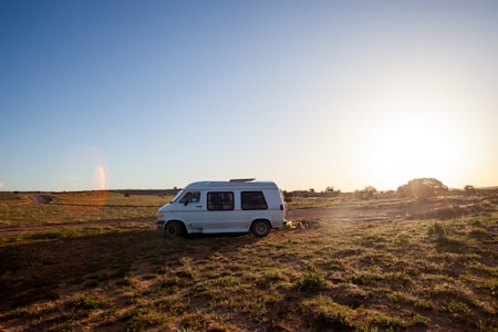 A camper van is parked on a wide-open field with sparse vegetation. The scene is illuminated by a setting or rising sun, casting a warm glow over the landscape. The sky is clear and expansive, adding to the sense of openness.