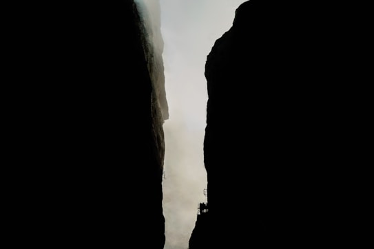 A narrow canyon or gorge with steep, dark rock faces on either side, separated by a thin strip of daylight. There are small, silhouetted structures or platforms visible on the sides of the rocks, giving a sense of scale and adding an element of human presence.