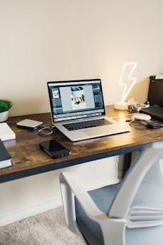 A modern workspace with a wooden desk featuring a laptop, smartphone, external hard drive, and a book. A chair is positioned next to the desk, and a neon lightning-shaped lamp is lit up on the right. A small plant in a pot adds a touch of greenery.