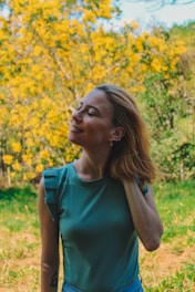 A woman wearing a teal sleeveless top stands in a lush garden with vibrant yellow flowers and greenery in the background. Her eyes are closed, and she has a serene smile on her face. The sunlight creates a warm and peaceful atmosphere.