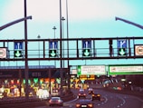 A highway scene featuring multiple lanes with cars heading towards toll booths. Overhead road signs with green arrows indicate open lanes, while electronic traffic signs display various directions and information. The scene is well-lit with artificial lighting, suggesting that it is either dusk or evening.