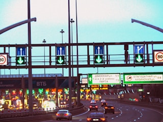 A highway scene featuring multiple lanes with cars heading towards toll booths. Overhead road signs with green arrows indicate open lanes, while electronic traffic signs display various directions and information. The scene is well-lit with artificial lighting, suggesting that it is either dusk or evening.
