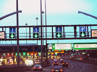 A highway scene featuring multiple lanes with cars heading towards toll booths. Overhead road signs with green arrows indicate open lanes, while electronic traffic signs display various directions and information. The scene is well-lit with artificial lighting, suggesting that it is either dusk or evening.