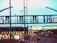 A highway scene featuring multiple lanes with cars heading towards toll booths. Overhead road signs with green arrows indicate open lanes, while electronic traffic signs display various directions and information. The scene is well-lit with artificial lighting, suggesting that it is either dusk or evening.
