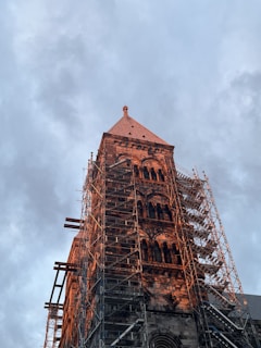 A historic stone tower under renovation is encased in scaffolding. The structure features intricate architectural details and is set against a backdrop of a cloudy sky, with soft lighting suggesting a time near sunrise or sunset.